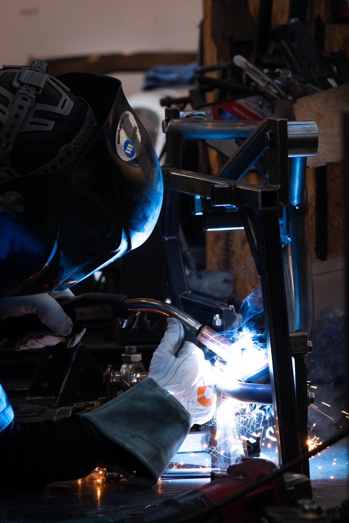 A welder in a black welding helmet and protective gloves generates bright blue-white sparks while working on a metal frame in a workshop setting.